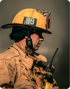 Side profile of a firefighter wearing a yellow helmet numbered 99 and holding a radio at a fire scene