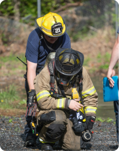 Two firefighters outdoors, with one in full gear crouching to check equipment while the other assists from behind