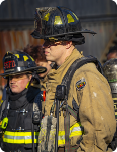 Two firefighters in turnout gear and helmets standing at an outdoor scene