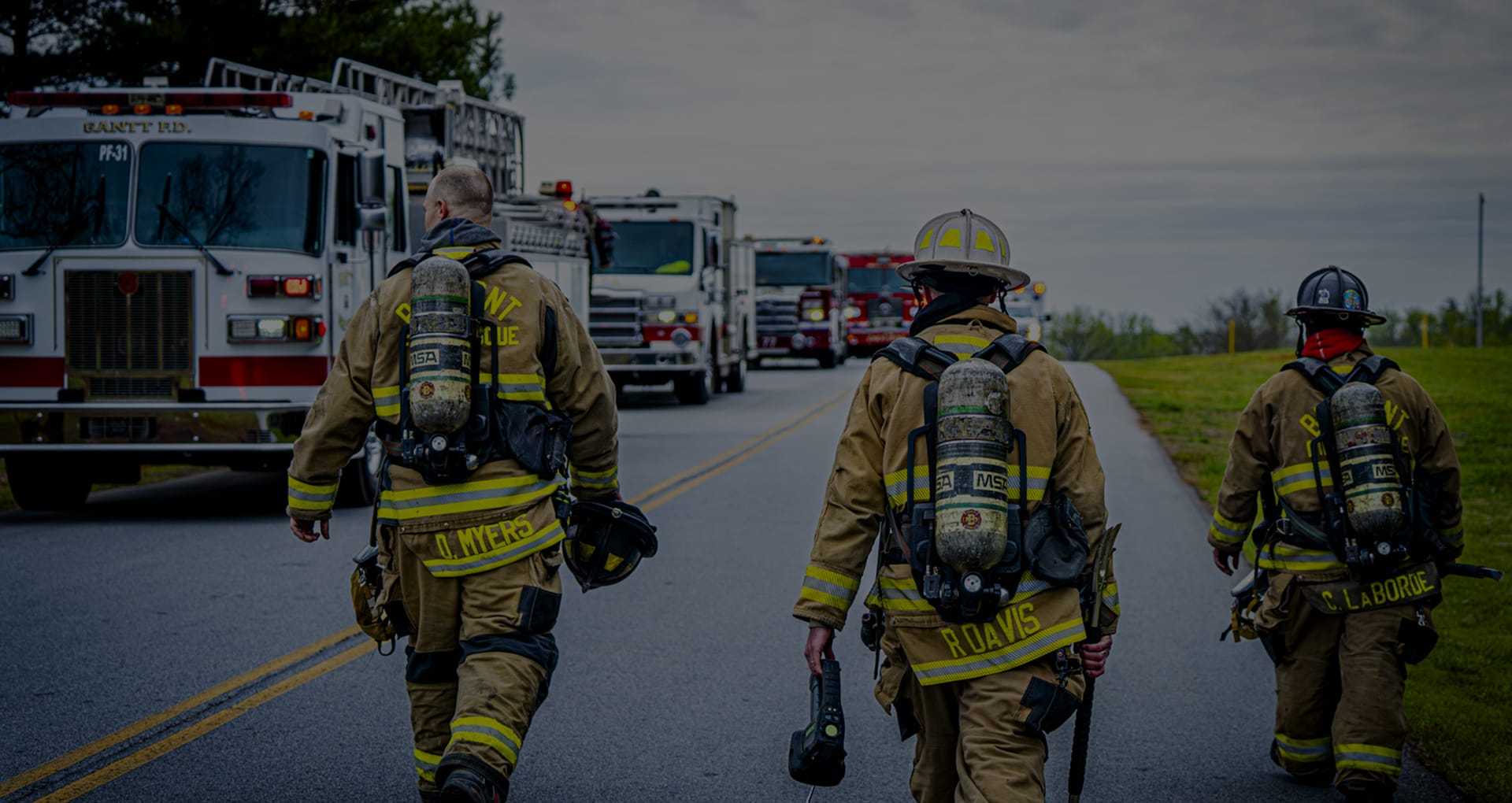Three firefighters in full gear walk down a road toward multiple fire engines lined up with flashing lights.
