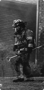 Firefighter in full gear with a breathing mask walks along a brick building carrying a hooked tool, with smoke drifting around the scene.