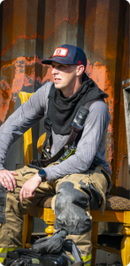 Firefighter in gear sits on a yellow chair in front of a rusted metal wall, wearing a cap with the number “13.”