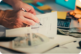 Person signing a document with a pen on a desk, with a calculator and U.S. dollar bills nearby.
