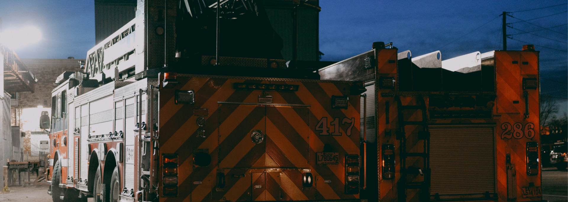 Two fire trucks parked at night, showing their rear chevron markings and equipment bays under a dark blue sky.
