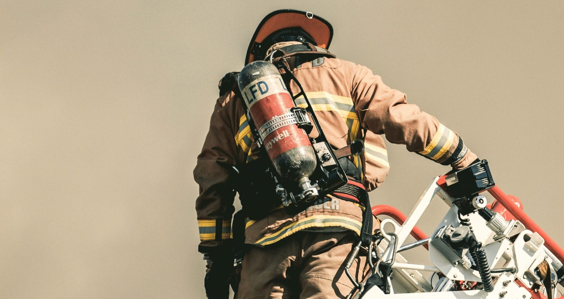 A firefighter in full turnout gear and SCBA tank climbs a ladder truck, viewed from behind against a neutral sky.