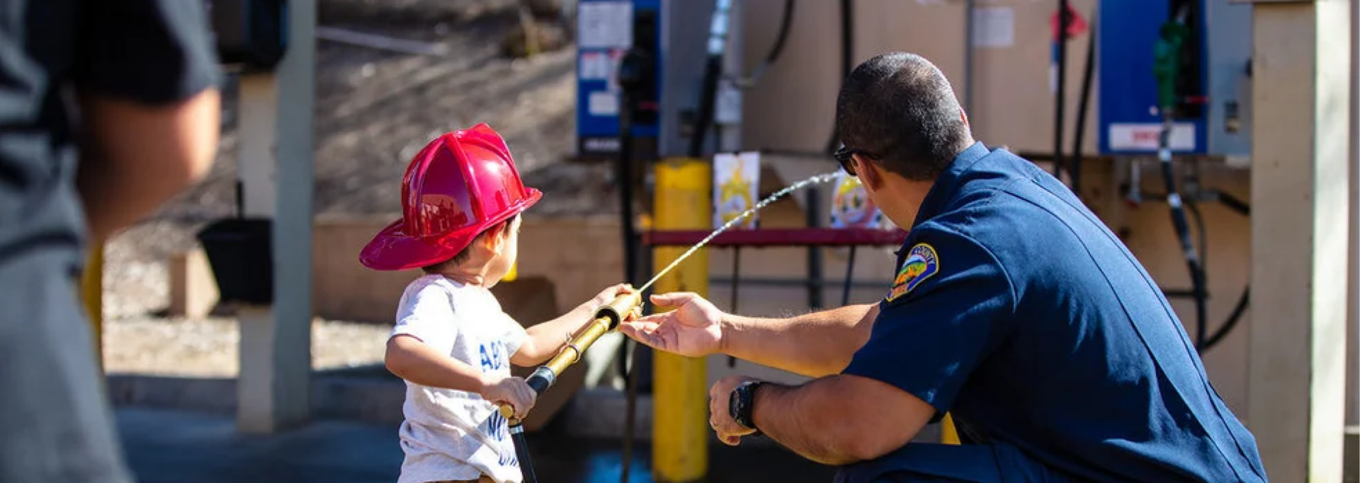 Firefighter guiding a young child wearing a toy helmet while holding a fire hose at a firehouse