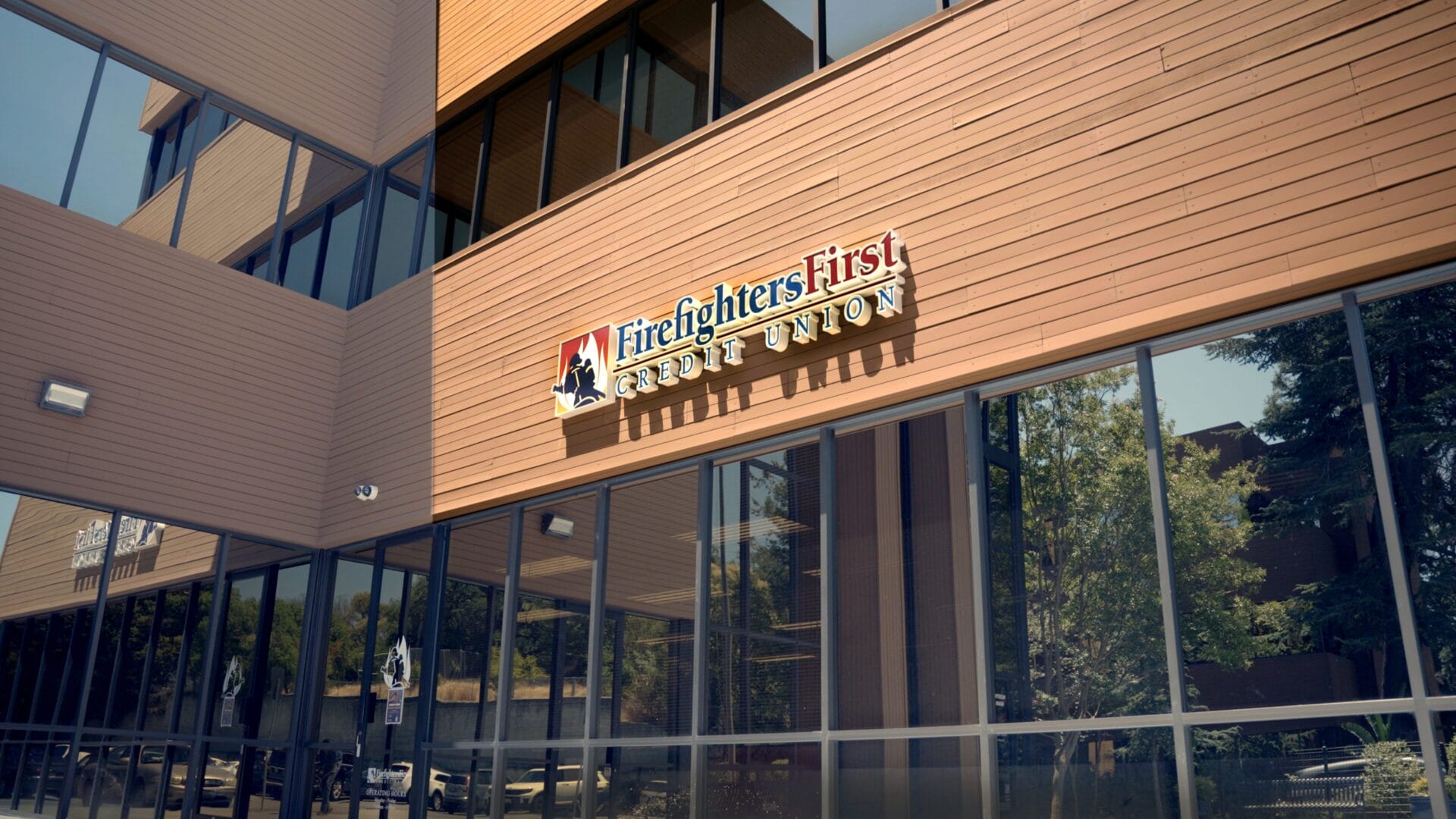 Exterior of the Firefighters First Credit Union Walnut Creek branch on a sunny day, with the FFCU logo and wordmark prominently displayed on a warm wood-panel facade above floor-to-ceiling glass windows reflecting nearby trees.