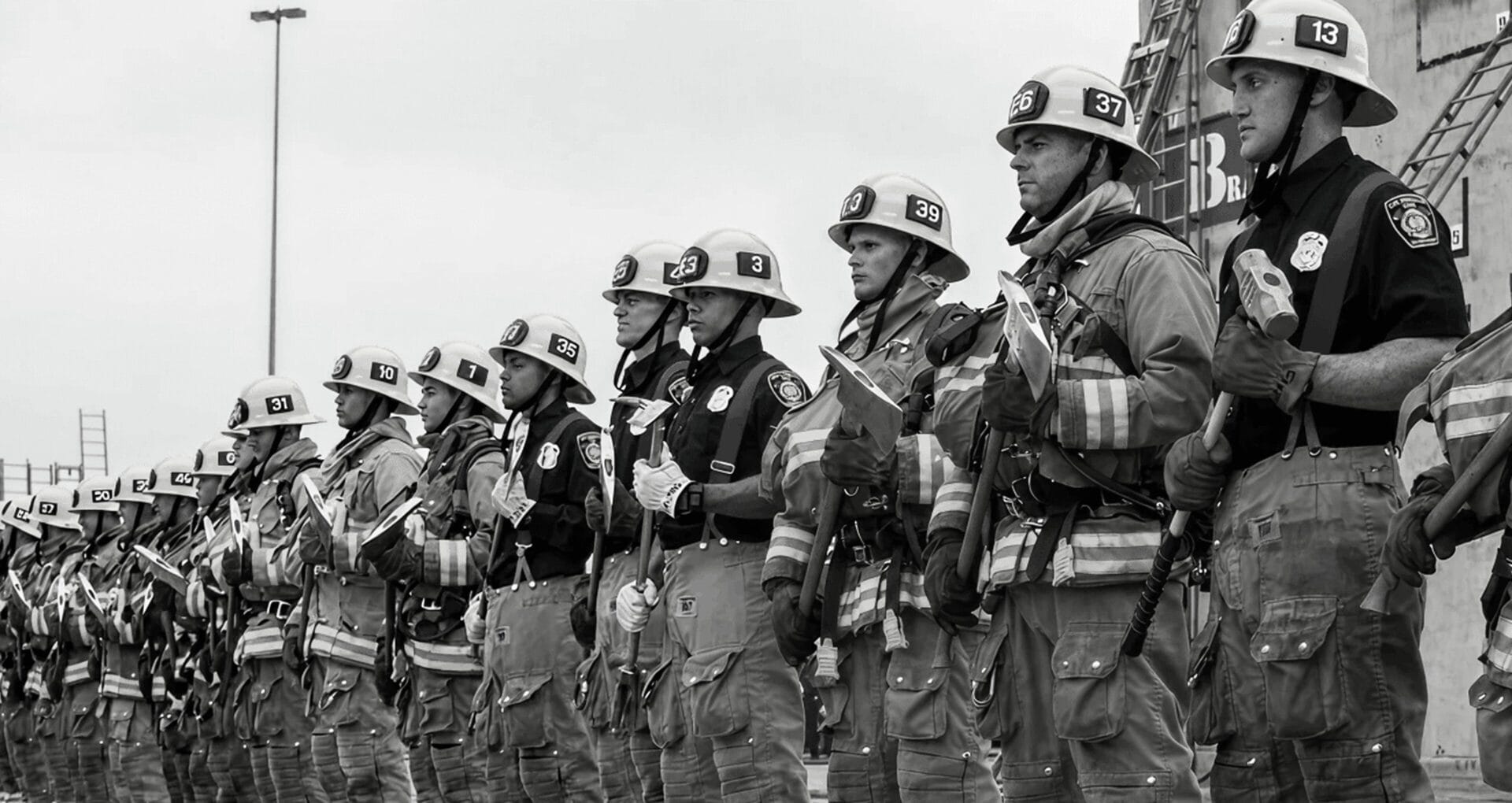 A line of firefighters in full turnout gear and numbered helmets standing at attention during a formal ceremony or graduation, photographed in black and white.