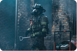 a firefighter in turnout gear and oxygen mask walking out of a burned brick building, heavy smoke visible.