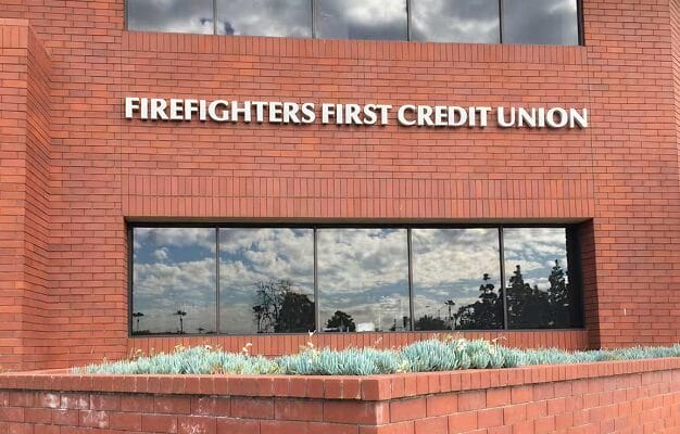 Exterior of a Firefighters First Credit Union branch, featuring a red brick building facade with the credit union name in large letters above a row of reflective windows and a raised brick planter with green shrubs.