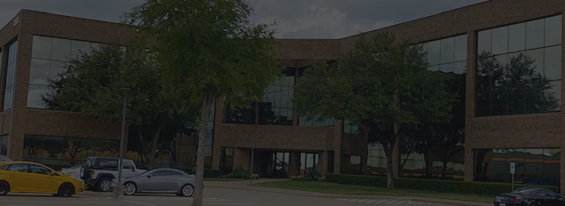 Exterior of the Firefighters First Credit Union Bedford branch, a two-story brick office building with large reflective windows and mature trees in the parking lot.